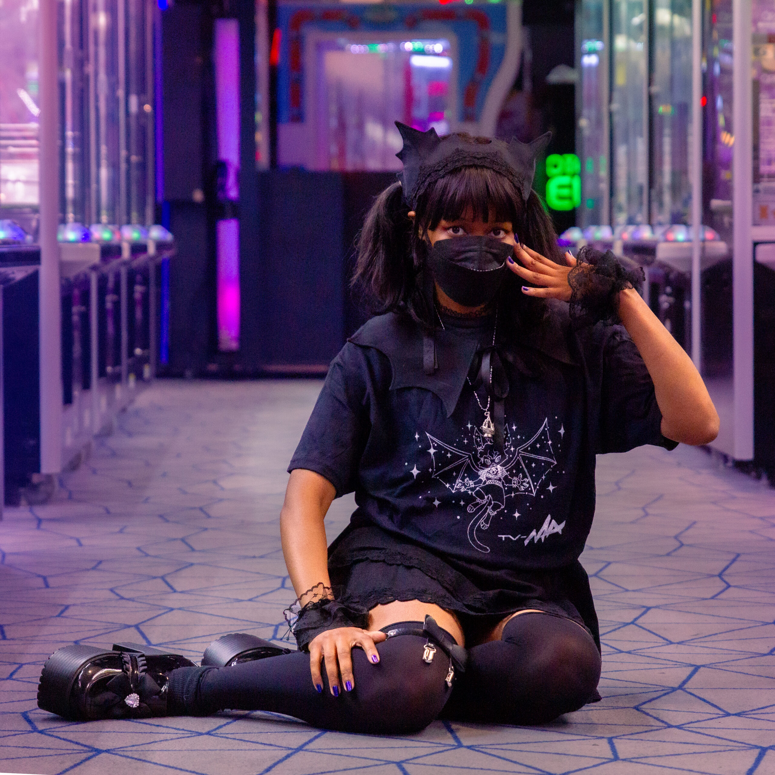 Lorraine Grate wearing cute, goth, kawaii fashion, wearing TV-MA Planets Will Spin Shirt in the middle of an arcade. She is sitting in a pose cutely, one hand spread in front of her face, wearing a KN95 mask. She is wearing a size L.